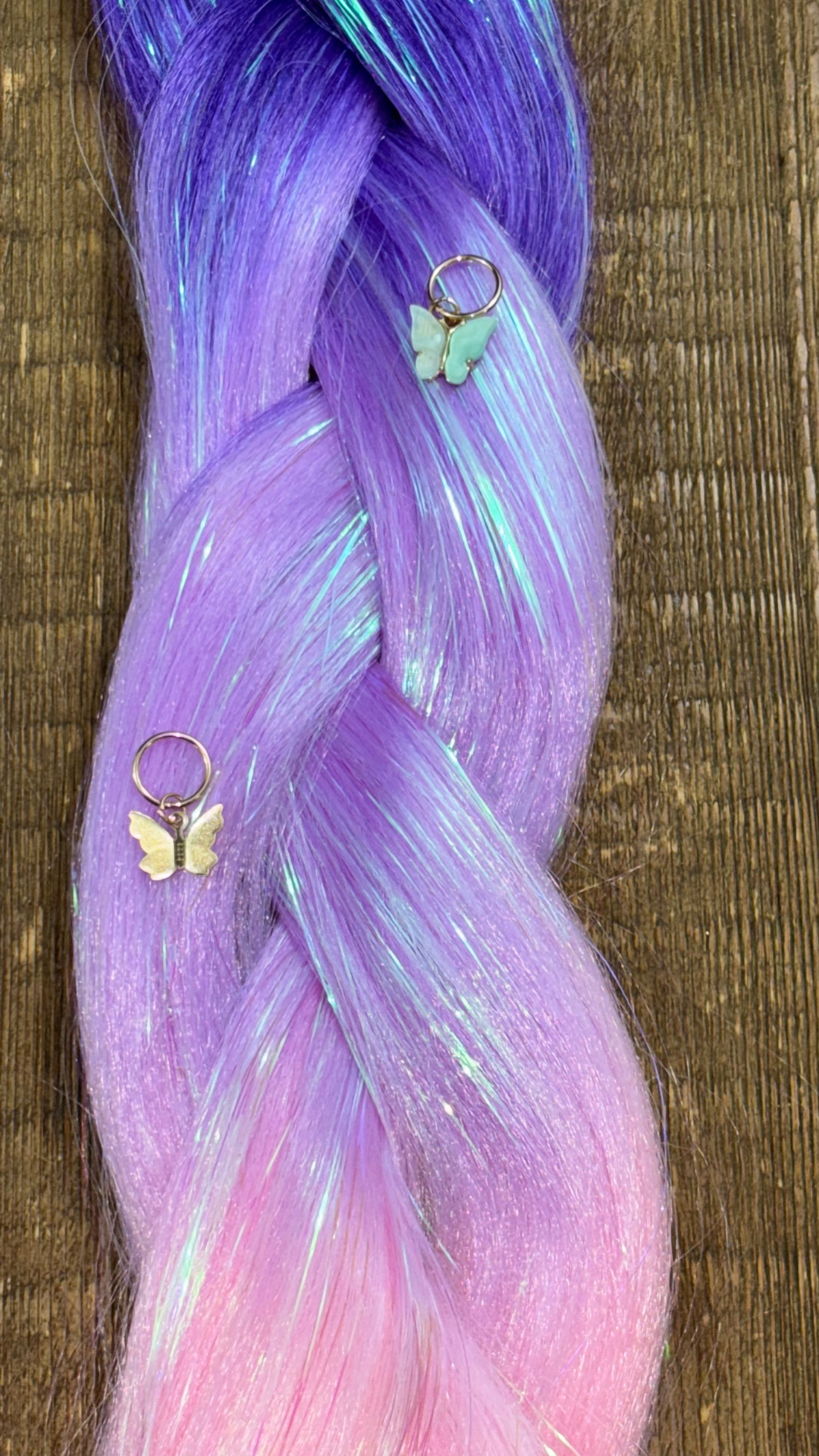 Close up shot of purple, lavender and pastel pink ombre girls hair extension braids with tinsel woven through and gold and pastel green butterfly hair charms on wood background.