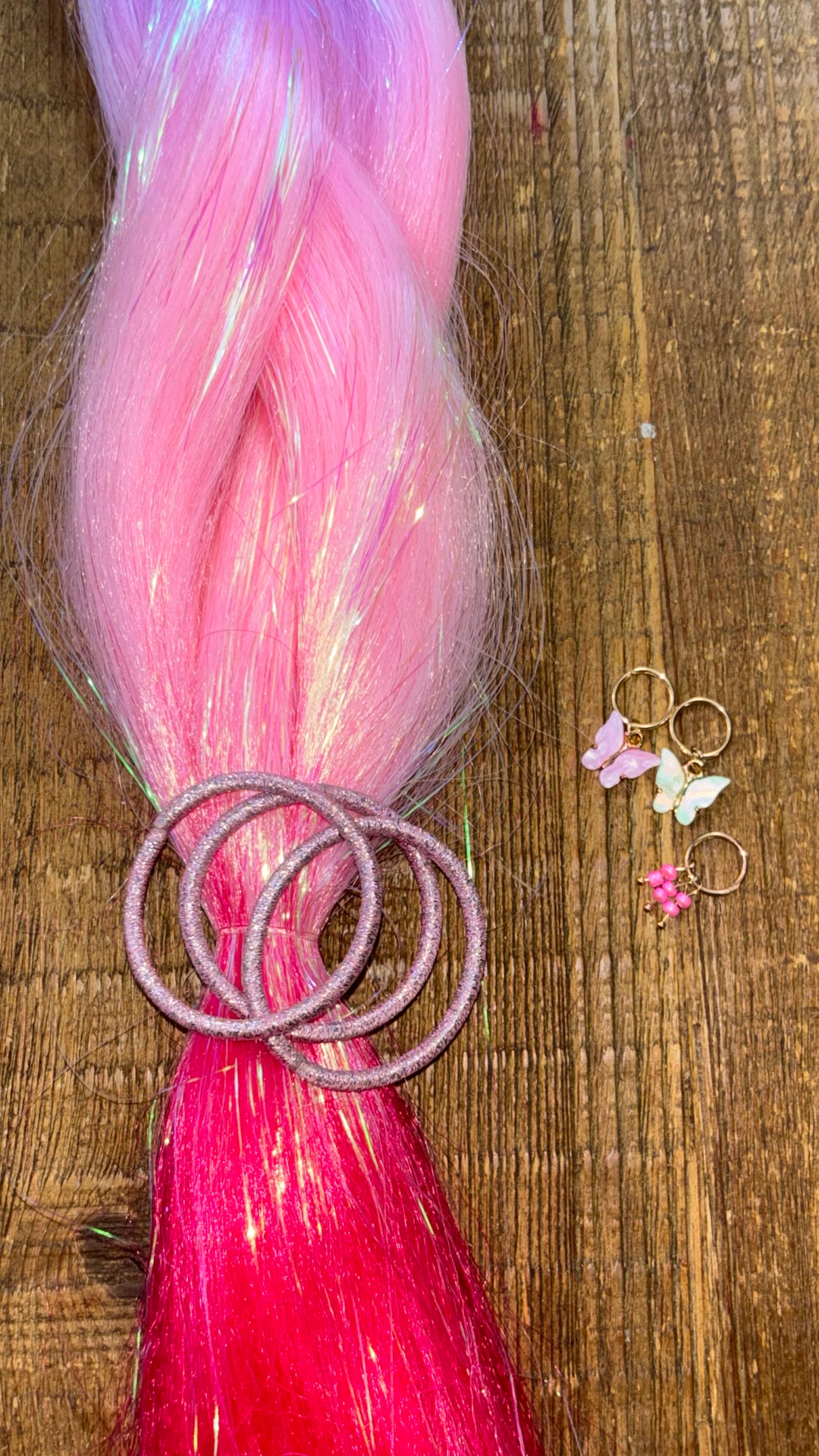 Close up of pastel and bright pink ombre girls hair braid extensions with tinsel woven through. Light pink metallic hair ties and pink and yellow butterfly hair charms on wood background.